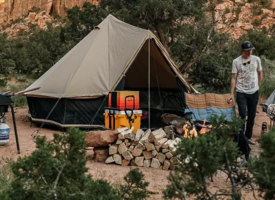 A man standing at a campsite nestled against rocky canyon walls, featuring a large tent, a neat pile of firewood, and a cooking station.