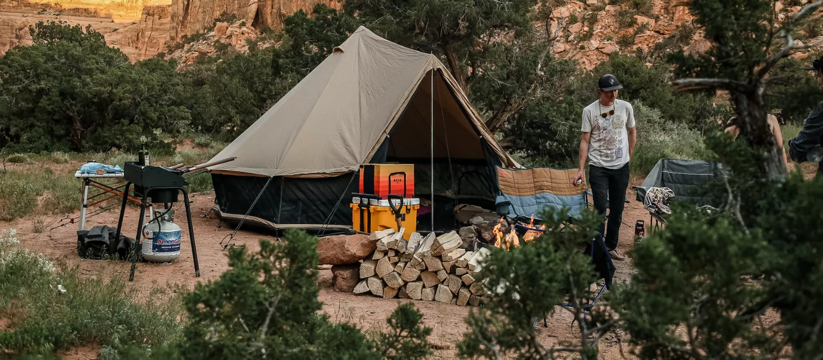 A man standing at a campsite nestled against rocky canyon walls, featuring a large tent, a neat pile of firewood, and a cooking station.