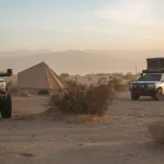 Full-time camping setup in desert with overland trucks, rooftop tents, and bell tent at sunset