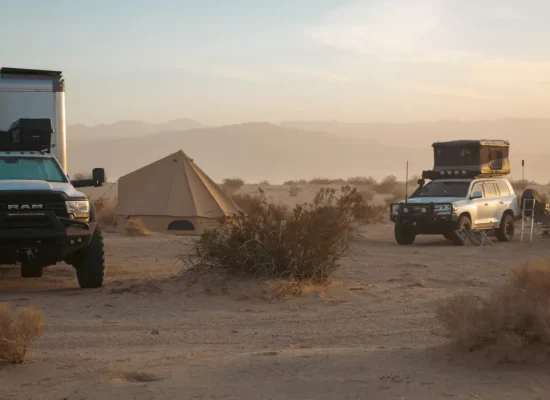 Full-time camping setup in desert with overland trucks, rooftop tents, and bell tent at sunset