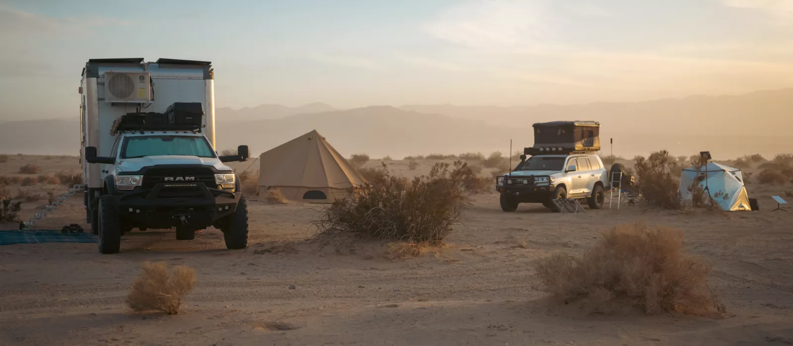 Full-time camping setup in desert with overland trucks, rooftop tents, and bell tent at sunset