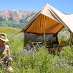 Spring glamping setup with canvas tent in blooming meadow and mountains in background
