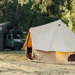 Spacious 6 person canvas camping tents set up at a wooded campsite with vehicles and outdoor gear nearby