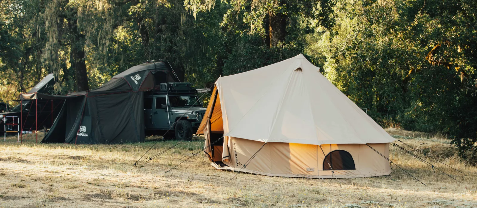Spacious 6 person canvas camping tents set up at a wooded campsite with vehicles and outdoor gear nearby