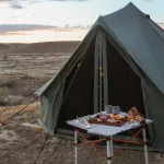 A green bell tent by white duck is set up for camping in a desert landscape during a vibrant sunset, featuring a folding picnic table with a charcuterie spread and string lights.