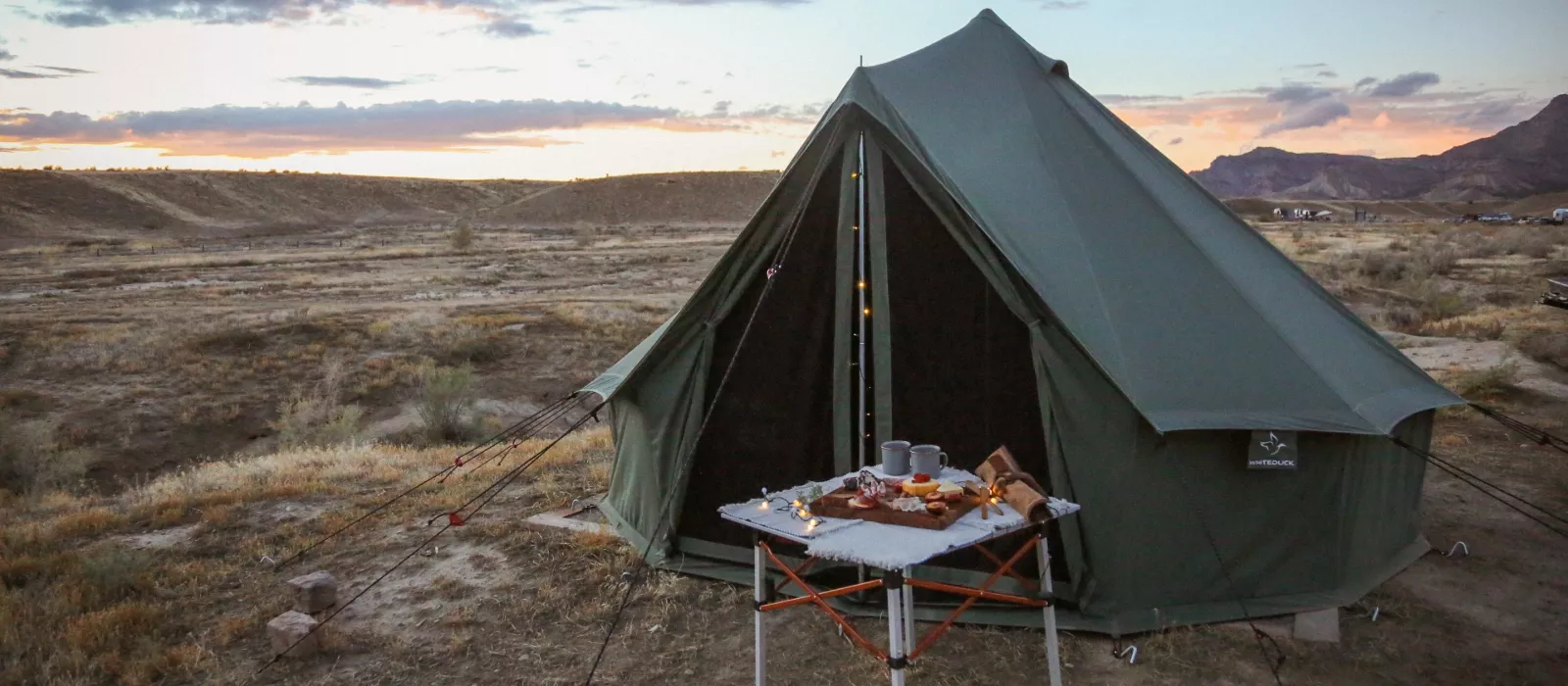 A green bell tent by white duck is set up for camping in a desert landscape during a vibrant sunset, featuring a folding picnic table with a charcuterie spread and string lights.