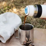 Cowboy coffee being poured from a thermos into a metal mug while camping outdoors