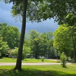 RV parked at a shaded campsite in Darien Lakes State Park with lush green trees, picnic area, and dramatic cloudy sky in the background