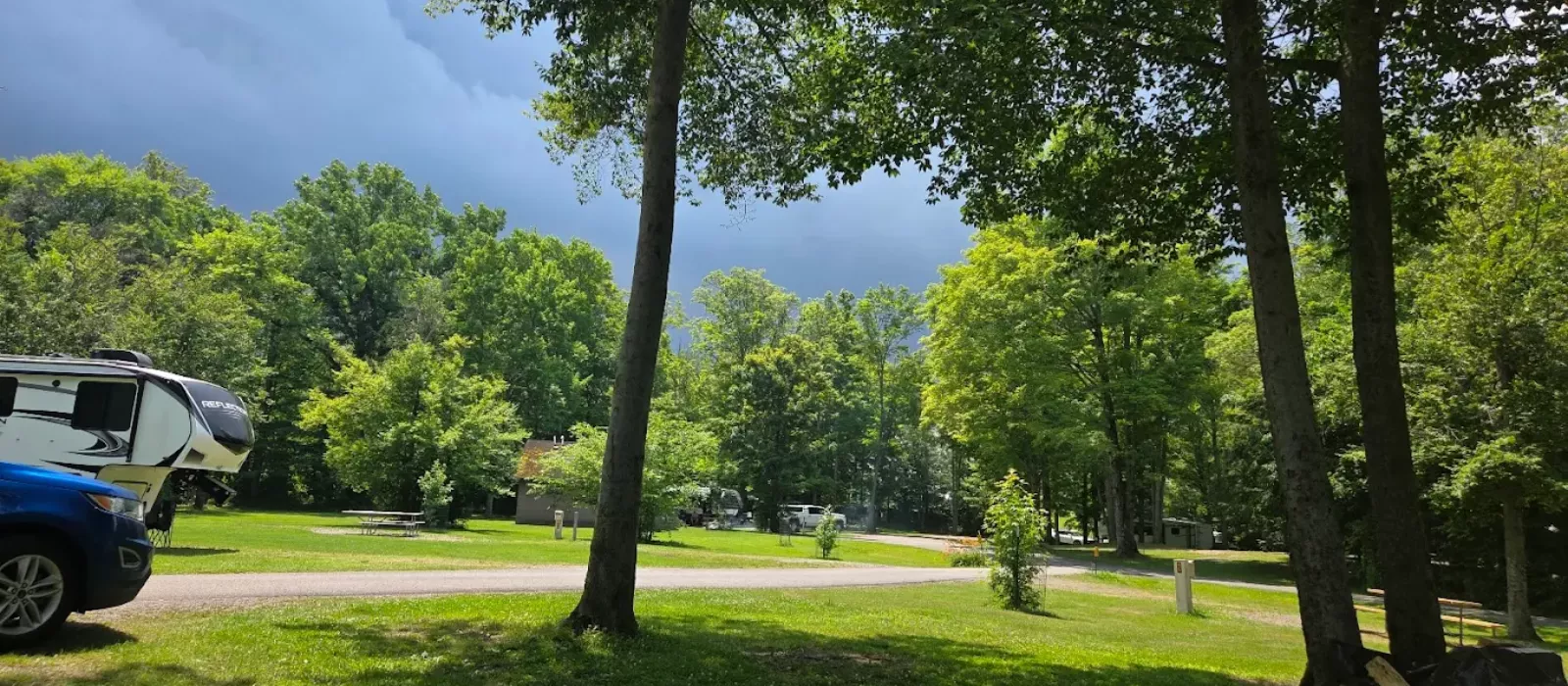 RV parked at a shaded campsite in Darien Lakes State Park with lush green trees, picnic area, and dramatic cloudy sky in the background
