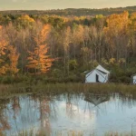 Luxury safari tents at Firelight Camps in Ithaca, nestled by a calm pond surrounded by vibrant autumn foliage. Credit: Firelight Camps GMB
