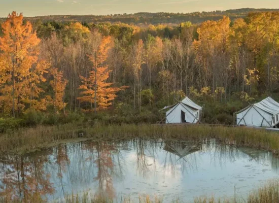 Luxury safari tents at Firelight Camps in Ithaca, nestled by a calm pond surrounded by vibrant autumn foliage. Credit: Firelight Camps GMB
