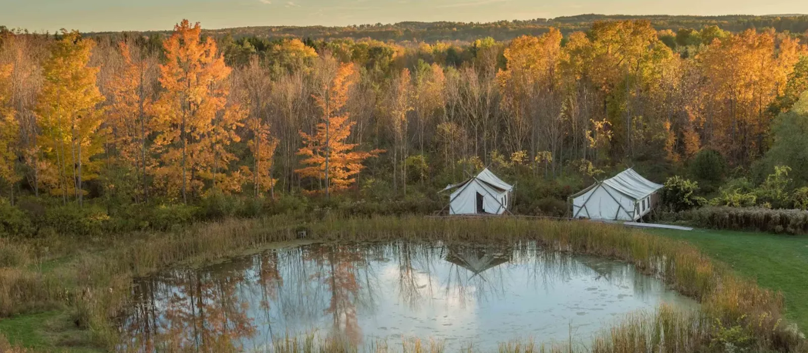 Luxury safari tents at Firelight Camps in Ithaca, nestled by a calm pond surrounded by vibrant autumn foliage. Credit: Firelight Camps GMB