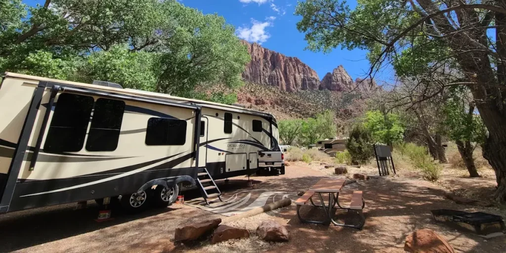 A large RV is parked at a campsite in Watchman Campground with a picnic table and dramatic view of the red rock cliffs of Zion National Park.
Credit: Watchman Campground GMB