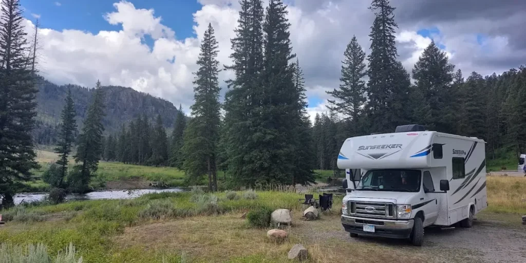 RV parked at Slough Creek Campground beside a flowing creek with pine trees, grassy meadow, and mountains under a cloudy sky.
Credit: Slough Creek Campground GMB