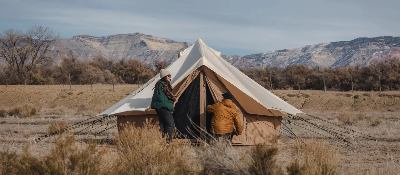 Two campers setting up a canvas bell tent in a dry mountain landscape during winter camping.
