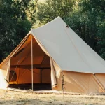 Canvas bell tent set up in a peaceful forest campsite with trees and dry grass surrounding it, sunlight filtering through the woods.