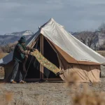 Person setting up a canvas bell tent in an open field with mountains and dry winter landscape in the background.