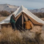 Campers setting up a canvas bell tent in a scenic mountain landscape during outdoor camping trip.