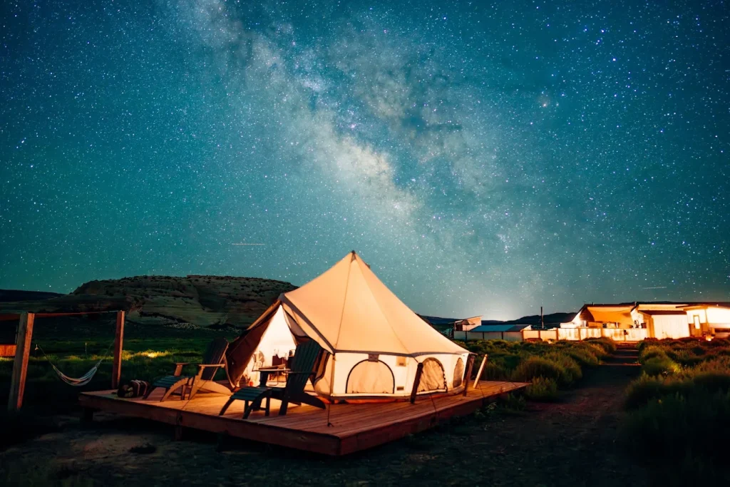 A glowing white bell tent sits on a wooden platform at a glamping site in Canyonlands, Utah, under a vibrant Milky Way galaxy and star-filled night sky.