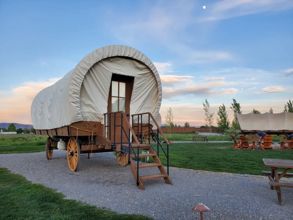 A modern Conestoga glamping wagon with a white canvas top and large wooden wheels parked on a gravel path at Conestoga Ranch.
