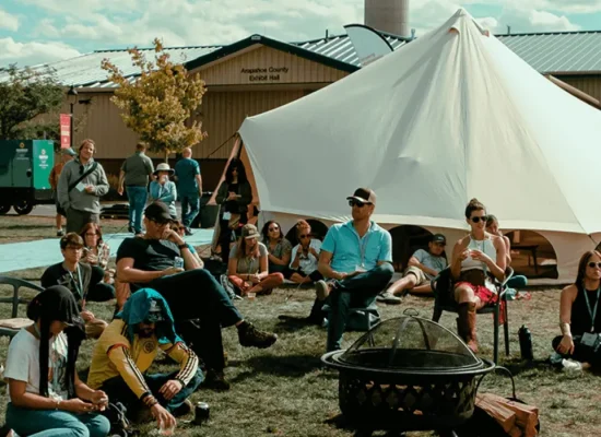 Optimus Avalon canvas tent displayed at an outdoor event with people gathered around the seating area and fire pit.