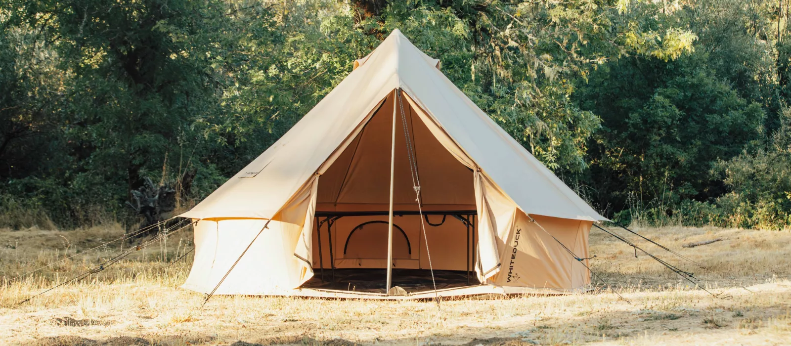 White Duck bell canvas tent set up at a forest campsite with open front entrance and guy lines secured outdoors.