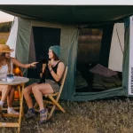 White Duck Outdoors 2026 Prota Canvas Tent set up in an open field with two campers relaxing under the awning during a camping trip.