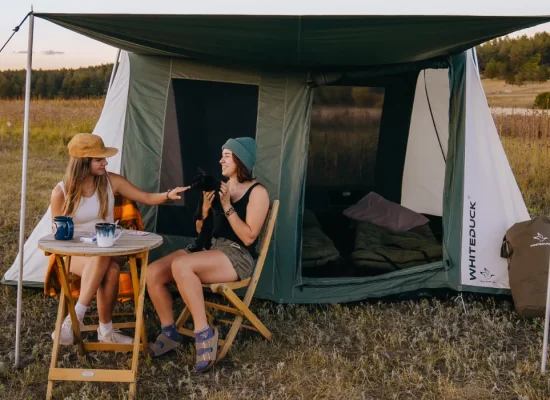White Duck Outdoors 2026 Prota Canvas Tent set up in an open field with two campers relaxing under the awning during a camping trip.