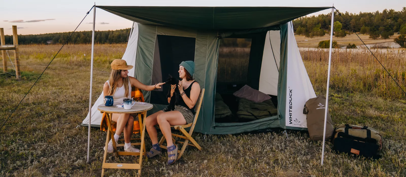 White Duck Outdoors 2026 Prota Canvas Tent set up in an open field with two campers relaxing under the awning during a camping trip.