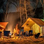 Canvas tents illuminated by a campfire during a nighttime camping trip in a wooded outdoor setting under a star-filled sky.