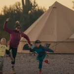 Family enjoying summer camping outside a canvas bell tent, with a parent and two children playing together at the campsite during sunset.