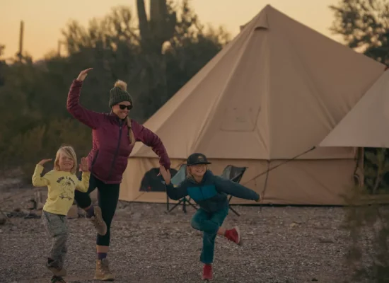 Family enjoying summer camping outside a canvas bell tent, with a parent and two children playing together at the campsite during sunset.