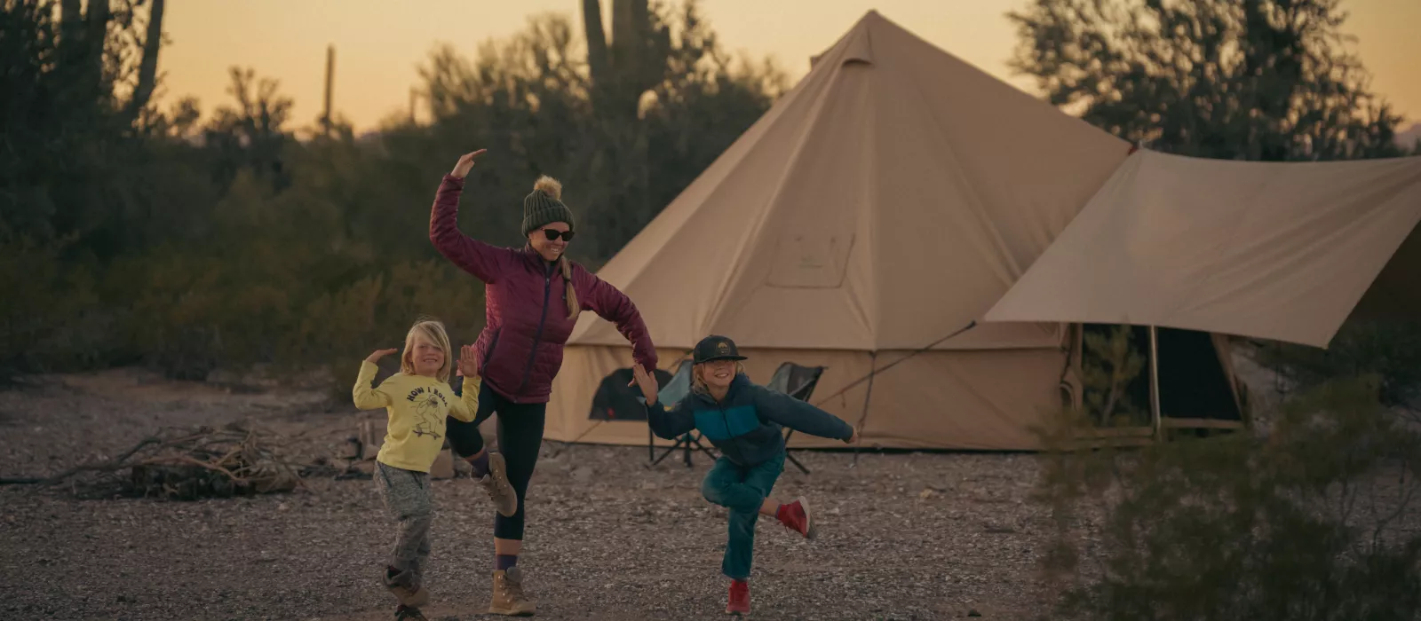 Family enjoying summer camping outside a canvas bell tent, with a parent and two children playing together at the campsite during sunset.