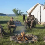 Family hunting camp beside a canvas tent near a lake, with parents and child gathered around a campfire, hunting gear, trucks, and a dog in an outdoor setting.