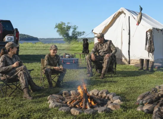 Family hunting camp beside a canvas tent near a lake, with parents and child gathered around a campfire, hunting gear, trucks, and a dog in an outdoor setting.