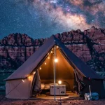 Canvas glamping tent illuminated at night beneath the Milky Way galaxy with red rock cliffs and a star-filled desert sky in the background.