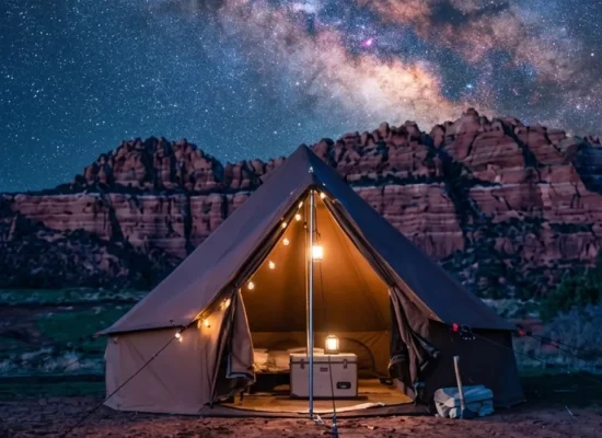 Canvas glamping tent illuminated at night beneath the Milky Way galaxy with red rock cliffs and a star-filled desert sky in the background.