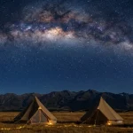 Canvas bell tents illuminated at night beneath the Milky Way galaxy, set against a mountain backdrop in a remote camping destination.