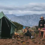 Two canvas tents set up at a mountain campsite with campers relaxing outdoors under a cloudy sky.