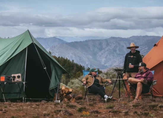 Two canvas tents set up at a mountain campsite with campers relaxing outdoors under a cloudy sky.