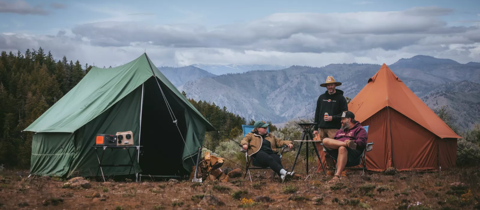 Two canvas tents set up at a mountain campsite with campers relaxing outdoors under a cloudy sky.