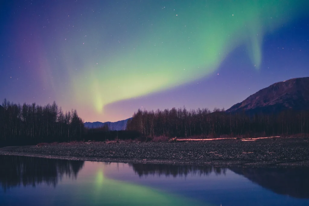 Northern Lights over a reflective lake with mountain and forest landscape under a starry night sky in Alaska.