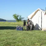 Hunter relaxing beside a canvas tent and hunting dog at a lakeside camp, preparing for the 2026 big game hunting season.
