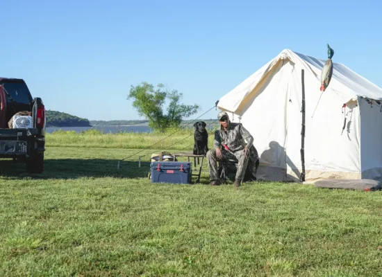 Hunter relaxing beside a canvas tent and hunting dog at a lakeside camp, preparing for the 2026 big game hunting season.