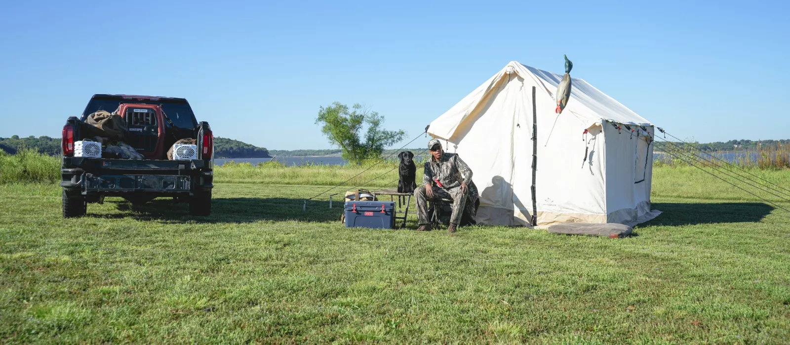 Hunter relaxing beside a canvas tent and hunting dog at a lakeside camp, preparing for the 2026 big game hunting season.