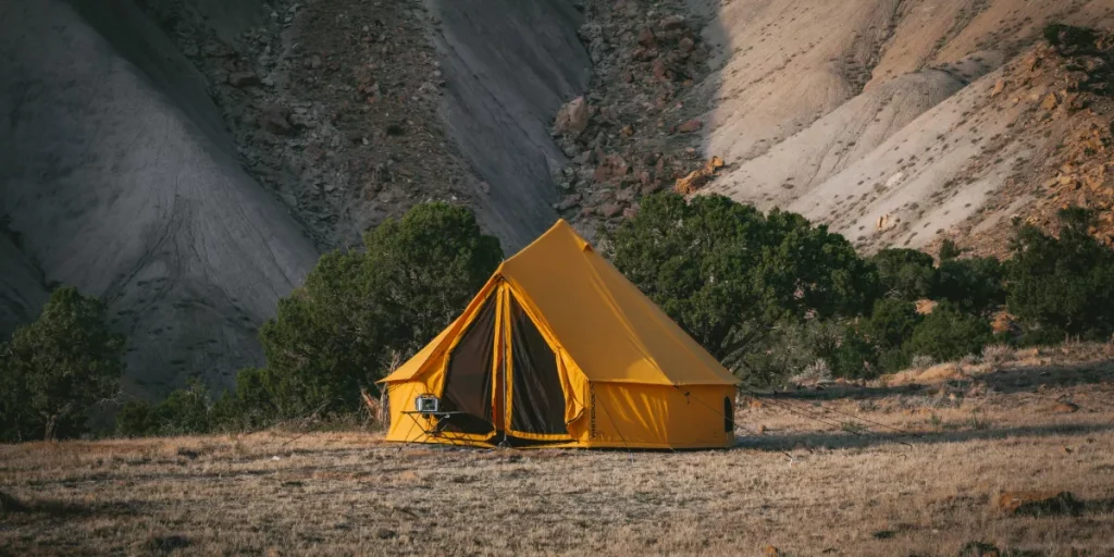Yellow bell tent pitched on a dry campsite with rocky mountain slopes and trees in the background, ideal for glamping and outdoor adventures.