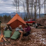 Campers gathered around a campfire with canvas tents, sleeping bags, a tarp shelter, and a trailer covered with a canvas tarp at a scenic woodland campsite