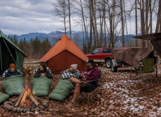 Campers gathered around a campfire with canvas tents, sleeping bags, a tarp shelter, and a trailer covered with a canvas tarp at a scenic woodland campsite