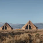 Four canvas bell tents set up in an open field with mountains in the background under a clear blue sky.