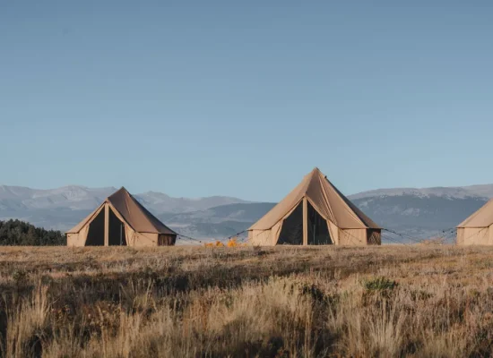 Four canvas bell tents set up in an open field with mountains in the background under a clear blue sky.